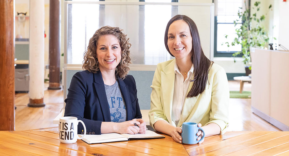 Alicia and Kristina sitting at a table, having coffee and smiling at the camera.